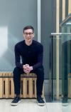 Kevin Poulter is sat on a bench crossing his hands, leaning forward and smiling at the camera. He has dark brown hair, wears small round glasses and is in all black clothing.