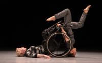 A female wheelchair-using dancer rests on her back, another female dancer balances on her head using the wheelchair for support