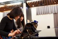 Two Asian woman looking at a musical score over a piano. The photo is shot from the side. The woman closest to the camera is wearing a black top, she has short brown hair with a fringe, and is holding a pencil. The woman next to her has long, dark brown hair with a fringe, and is wearing a grey top.