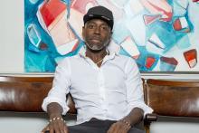 Headshot of Kevin Osborne. He is a Black man wearing a white shirt, black trousers, and a black cap. He sits on a brown chair in front of a painting.