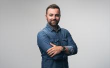 Liam McMullan stands in front of a grey background with his arms folded