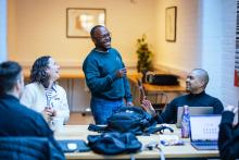 A Black man is standing in a workspace room, laughing with four other people who are sat down. They all have laptops and there are water bottles, mugs, and bags on the desk in front of them.