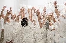 Image of people dressed in white, hands aloft, with white confetti/petals falling