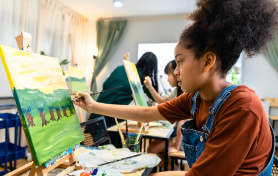 A school pupil takes part in an art class. she is painting a landscape with trees on a canvas