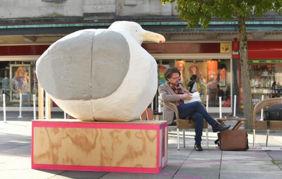 Statue of a large seagull with a man sitting crossed legged on a bench reading in the background