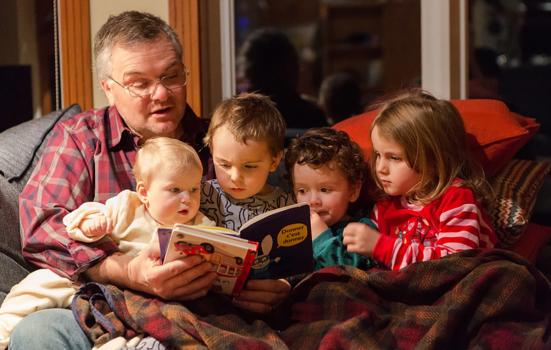 Photo shows a grandfather reading bedtime stories to three toddlers and a baby on a sofa