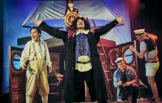 A pirate on stage performing Treasure Island in front of a ship prop and his crew