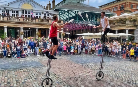 Two unicyclists surrounded by spectators juggle with clubs in front of Covent Garden Piazza