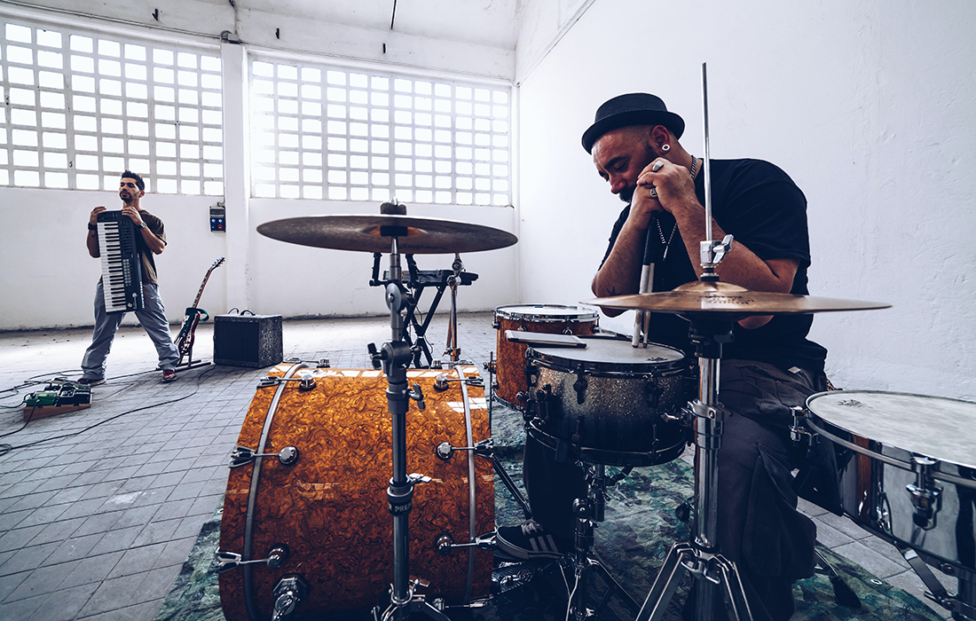 a man sitting behind a drum-kit in the foreground and a man standing in the background with a keyboard