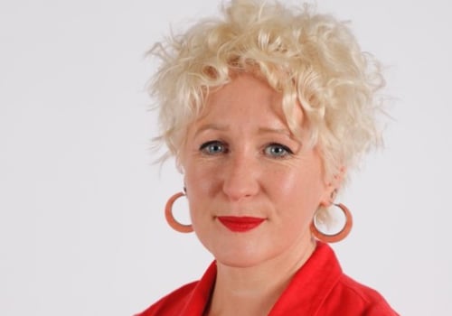 Rachel Dickson smiling. She has short platinum blonde curly hair with round orange hoop earrings. She is wearing red lipstick and a red shirt.