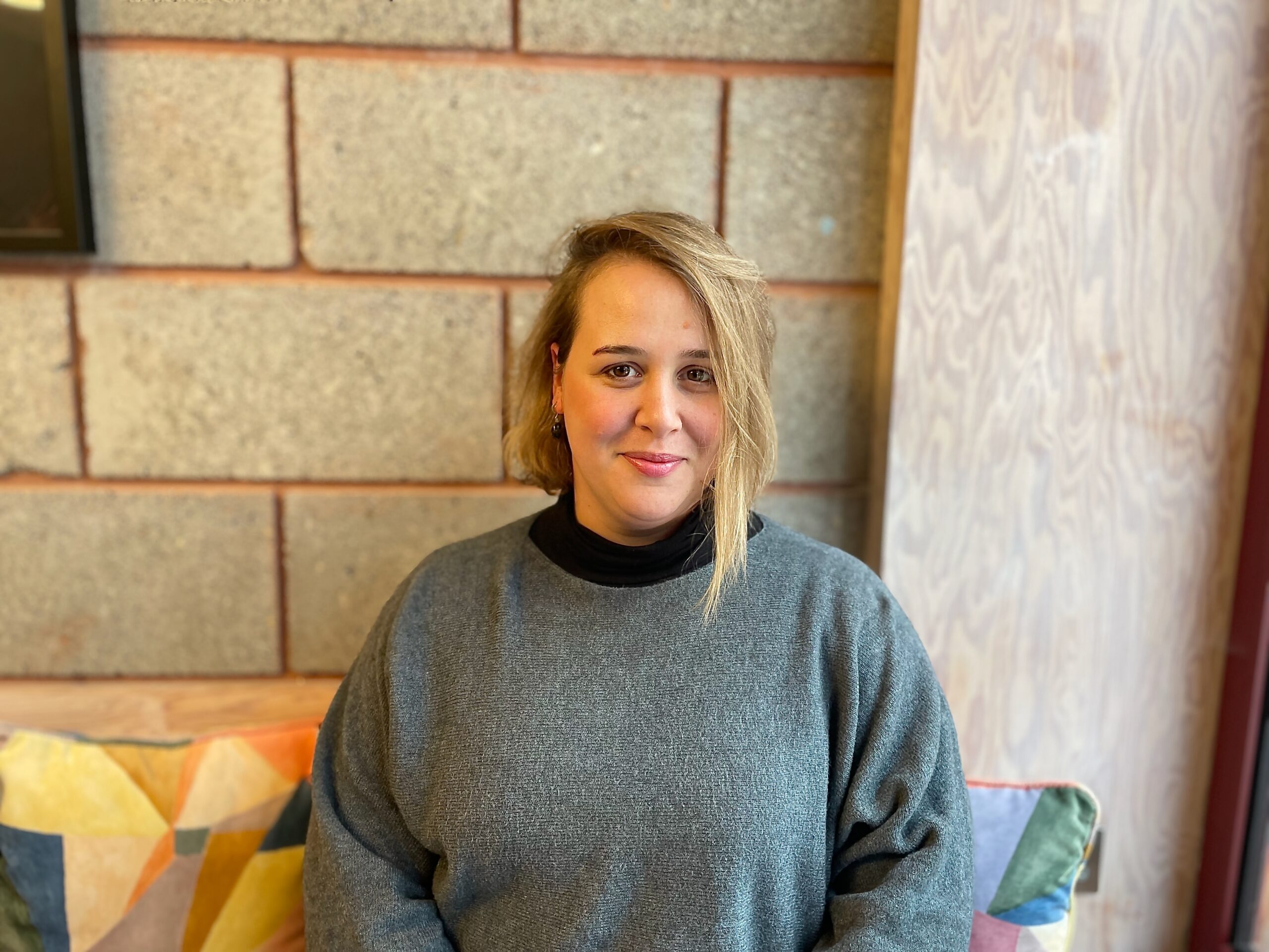 Rowan Rutter is sat down and smiling at the camera. She had blonde hair and is wearing a grey jumper with a black turtleneck underneath