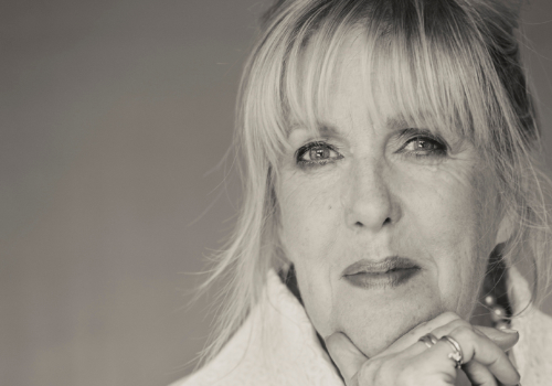 Black and white headshot of Sandra Gunn. She is positioned to the right of the image. Gunn has blonde hair with a fringe and is looking into the camera. She is holding her fingers to her chin.