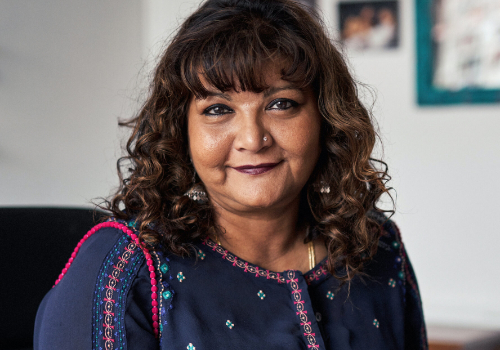 Tanika Gupta is an Asian women with curly, short brown hair and a full fringe. She is smiling at the camera. She has a nose piercing and is wearing purple lipstick.