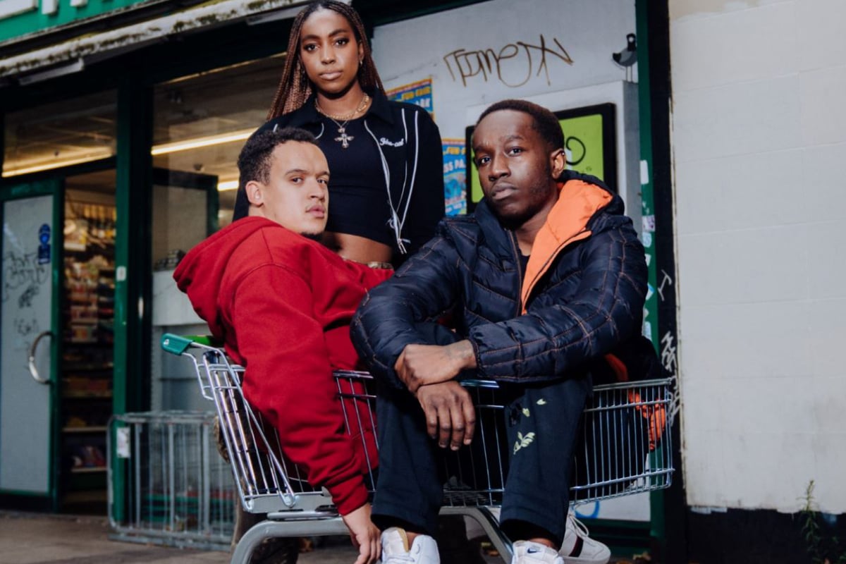 National Youth Theatre's production shot from 'GONE TOO FAR!'. There are two young men sitting in a trolley and a young women standing behind; they are all looking into the camera. Corner shop in the background.