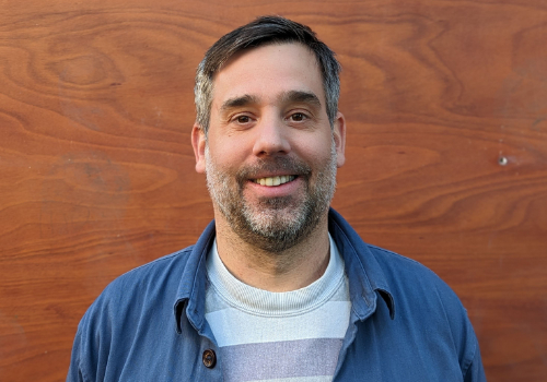 Man who has brown/grey hair and a brown/grey beard. He is smiling at the camera. He is stood against a wooden wall. He wears a blue shirt with a striped white + grey t-shirt underneath.