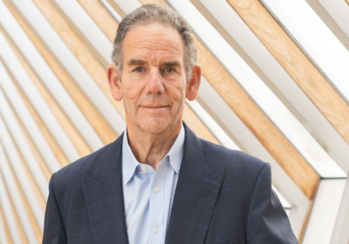 Christopher Rodrigues CBE in the The Royal Ballet School 2022. He is a man wearing a suit, smiling at the camera with a diagonally-designed, wooden/metal wall behind him.