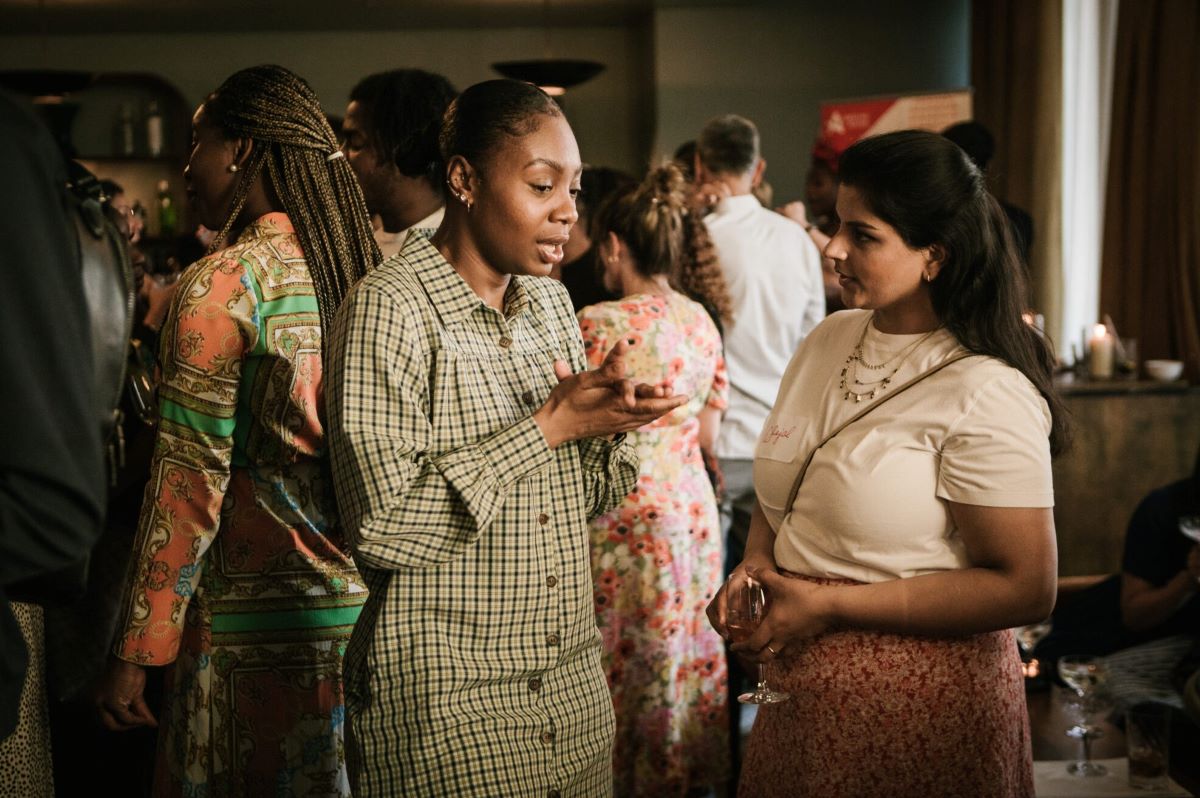 A Black woman speaking to a mixed-race woman at a social event. The Black woman is wearing a tartan dress with her hair in a tight bun, while the mixed-race woman wears a white top & skirt and has her hair down.