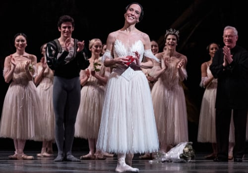 Laura Morera is a white woman. She is front centre stage in a long, white dress. Her brown hair is tied in a bun. She is wearing ballet shoes. The other performers are behind her, applauding her.