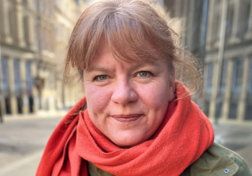 Amanda Smethurst is a white woman. She stands outside, smiling at the camera. There are buildings in the background (blurred). She has strawberry blonde hair, green eyes and is wearing taupe lipstick. She wears a coral coloured scarf and a green coat. Her hair is up, but she has a fringe which covers her forehead.