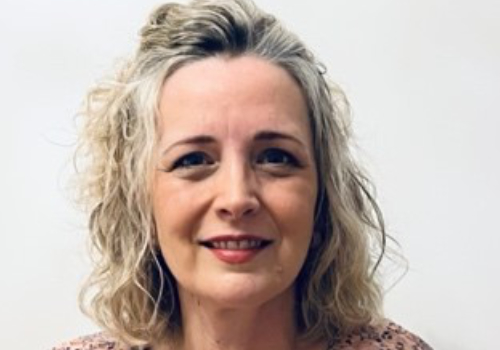 Tedbury is a white woman who has silver and blonde, curly hair that reaches her shoulders. She is wearing red lipstick. She is looking at the camera, smiling against a white background.