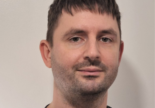 Wright is a white man pictured against a white wall. He has short, brown hair and brown eyes with a slight fringe falling over his forehead. He also has short stubble across his upper lip and chin, and is looking directly at the camera.