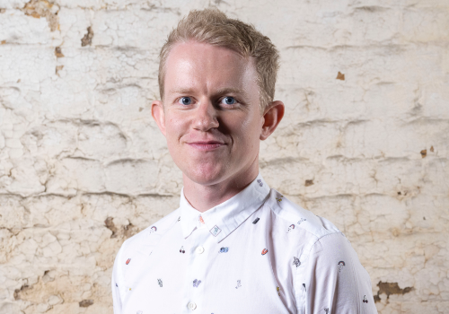 Harry Hickmore, Development Director, Orchestra of the Age of Enlightenment (OAE). He is a white man with blonde hair, standing against a white, brick wall. He is wearing a white shirt with small patterns, and smiling at the camera.