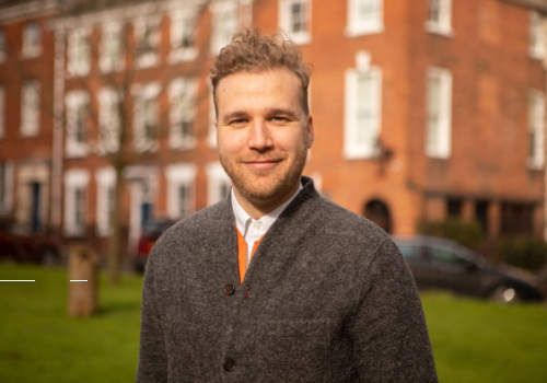 Jon Davis, Director of Cement Fields. He is a white man who has strawberry blonde hair, and is looking at the camera smiling. He wears a grey coat, with a white shirt underneath. The background is blurred, though there is a building with many windows.