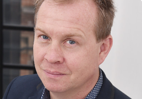 James Stirling, outgoing BBC Music Executive Editor. He is a white man with strawberry blonde hair and green eyes. He wears a blue suit and polka dot, blue and white shirt.