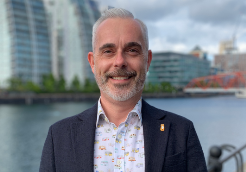 Darren Grice, Director of Culture, Salford City Council (SCC) and The Lowry. He is a white man with grey hair, a moustache and stubble. He wears a black blazer and patterned white shirt. In the background, there is a body of water and buildings which are blurred. He is the focus of the image.