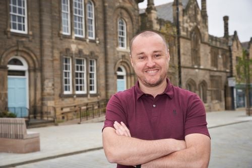 Stuart Egan, incoming Managing Director of The Exchange. Egan is a 43-year old man. He is photographed standing in front of The Exchange with his arms folded. He is smiling, wearing a grape-coloured polo shirt.