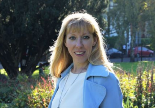 Emma Flatley, Director at Theatr Brycheiniog. A white woman with blonde hair and a full fringe, she wears a white top, blue blazer, and silver necklace. She is photographed outside in a park, the sun is shining on her.