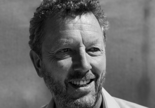 Paul Crewes, incoming Chief Executive, York Theatre Royal. A black and white image captures Crewes from the shoulders up. He is angled slightly to the side, smiling while not making eye contact with the camera. He is a white middle-aged man with curly hair, a trimmed moustache and beard, and is wearing a shirt.