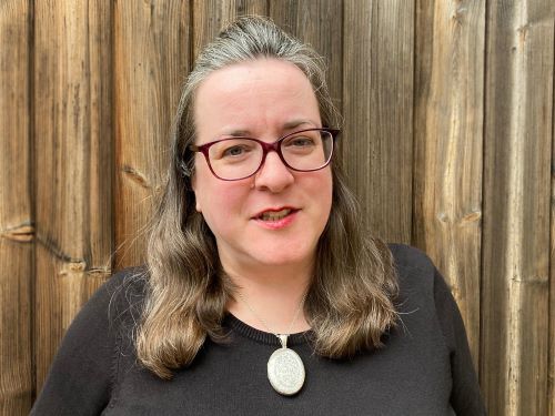 Julia Bird, incoming Executive Director, Poet in the City. She is photographed against a wooden fence, wearing a black top, silver necklace and maroon glasses. She has light brown-grey hair.