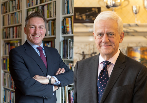 (L to R) Simon Larter-Evans and Stefan Anderson. Larter-Evans is positioned in a room with a bookshelf in the background, standing with his arms crossed. He is dressed in a navy suit, sporting a plum tie, and a warm smile. Anderson, an elderly gentleman with grey hair, stands in a grand room painted in yellow. He is attired in a grey suit complemented by a blue tie with polka dots.