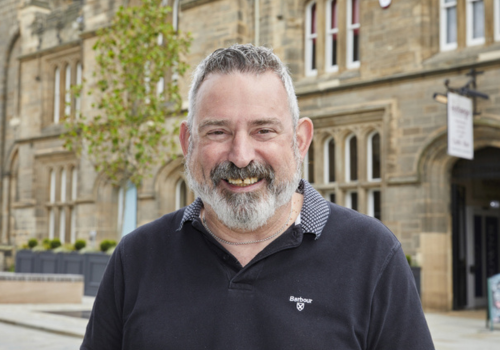 Adam Langman, photographed stood outside Exchange 1856. He has grey hair and wears a black polo top.