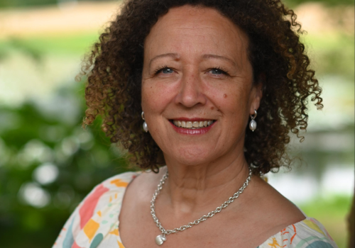 Julie Ellen is photographed outside. The background is blurred. She has curly, brown hair and wears silver jewellery.